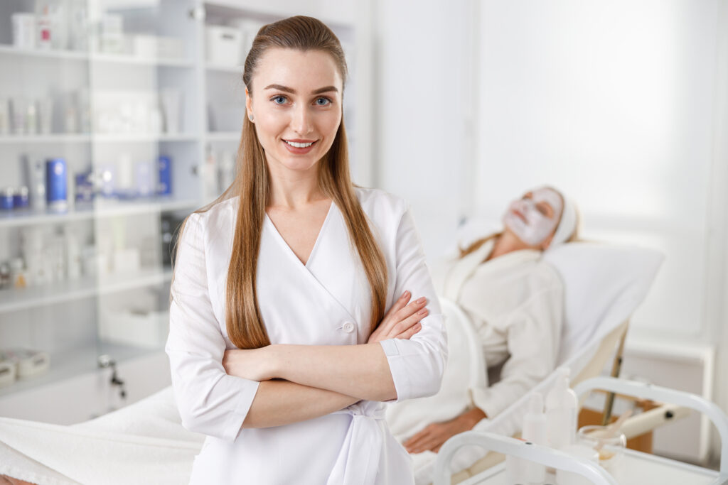 Lekarka medycyny estetycznej w gabinecie, stojąca obok leżaka, na którym leży pacjentka z nałożoną maską na twarzy. 