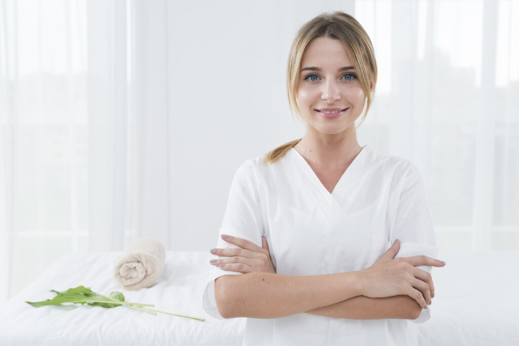 Techniczka usług kosmetycznych w białym kitlu, stojąca przed łóżkiem, na którym jest zwinięty ręcznik.